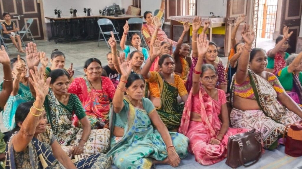 Women sitting on the floor, evidently in a meeting, some of whom are raising their hands in approval.